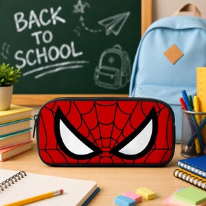 May include: A rectangular pencil case featuring a vibrant red background with a black spiderweb pattern and two large, white, angular eye shapes. The case has a black zipper closure along the top edge and a structured, rounded design for storage.