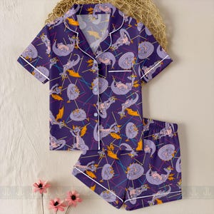 May include: Purple pajama set featuring a repeating pattern of a cartoon dragon character. The short-sleeved top and shorts have white trim and buttons. The dragon is purple with orange wings and is set against a background of colorful lines.