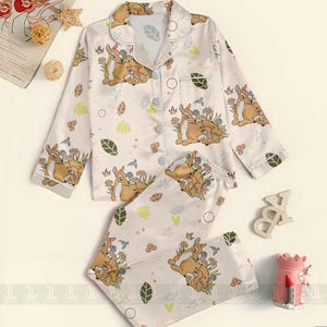 Op de afbeelding: Cr&egrave;mekleurige pyjamaset met een Bambi- en Stampertje-print. De top met lange mouwen heeft een knoopsluiting en een reverskraag. De broek heeft een elastische tailleband. De set bevat een herhalend patroon van Bambi, Stampertje, bloemen en bladeren.