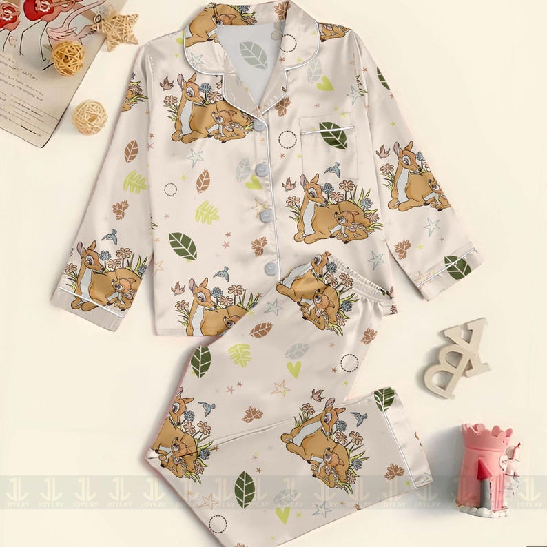 Op de afbeelding: Cr&egrave;mekleurige pyjamaset met een Bambi- en Stampertje-print. De top met lange mouwen heeft een knoopsluiting en een reverskraag. De broek heeft een elastische tailleband. De set bevat een herhalend patroon van Bambi, Stampertje, bloemen en bladeren.
