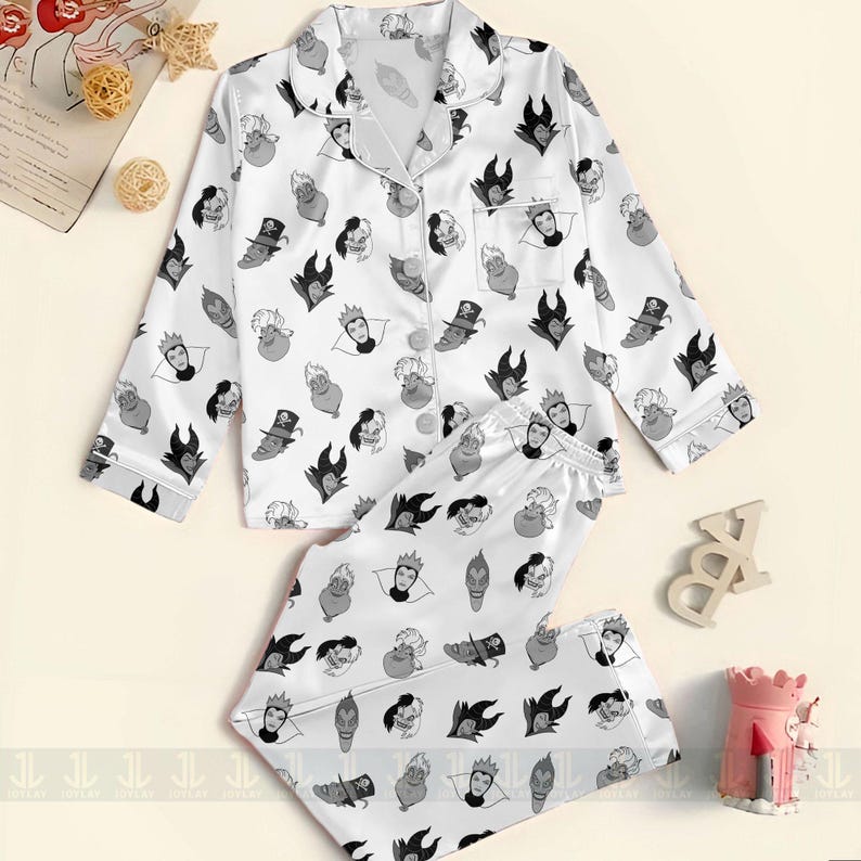 K&ouml;nnte beinhalten: Wei&szlig;es Satin-Pyjama-Set mit einem sich wiederholenden Muster von Cartoon-B&ouml;sewicht-Gesichtern in Schwarz und Grau. Das Set beinhaltet ein Button-Down-Oberteil mit Reverskragen und langen &Auml;rmeln sowie eine passende Hose mit Kordelzug. Die Pyjamas sind eine bequeme Schlafbekleidung.