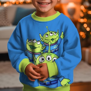 May include: A blue sweatshirt featuring three green cartoon aliens with large eyes. The sweatshirt has a green collar and cuffs. The child is wearing the sweatshirt. The background is blurred, with a Christmas tree and lights visible.