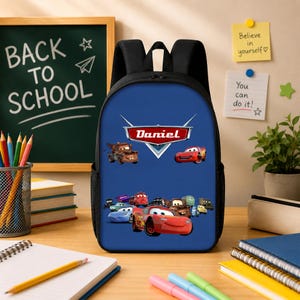 May include: A blue backpack featuring a graphic print of characters from the movie Cars, with the name Daniel displayed in a red and white logo at the top. The backpack has black straps and side mesh pockets, centered on a desk with school supplies.