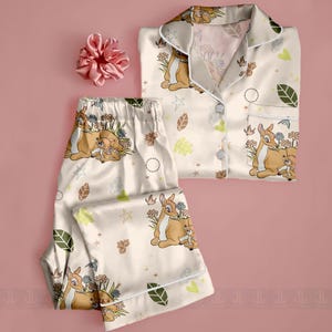Op de afbeelding: Cr&egrave;mekleurige pyjamaset met een Bambi en Stampertje print. De set bevat een overhemd met knopen en een zak en een broek met een elastische tailleband. Een bijpassende roze scrunchie is ook inbegrepen. De pyjama's zijn versierd met bloemen en bladeren.