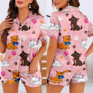 May include: Pink satin pajama set featuring a pattern of cartoon cats, paw prints, and clouds. The short-sleeved top and shorts have white trim. The cats are orange, brown, and white, with blue and red bows. The set is designed for comfort and style.