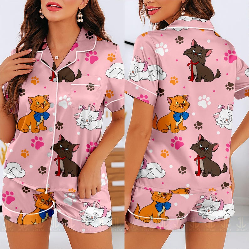 May include: Pink satin pajama set featuring a pattern of cartoon cats, paw prints, and clouds. The short-sleeved top and shorts have white trim. The cats are orange, brown, and white, with blue and red bows. The set is designed for comfort and style.