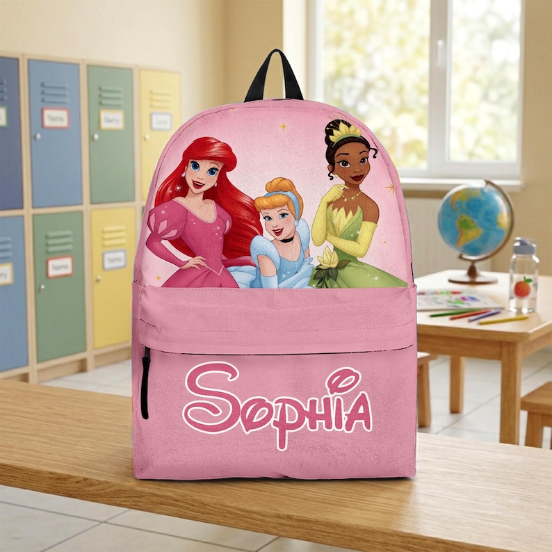 May include: A pink backpack featuring a graphic print of three animated princesses with red, blonde, and dark hair. The front pocket displays the name Sophia in a white, stylised, cursive font. The bag has a black top handle and black zipper pulls.