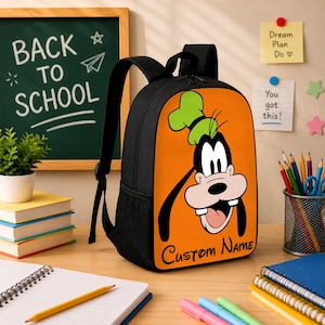 Peut inclure: Un sac &agrave; dos noir avec un panneau avant orange orn&eacute; d'une illustration du personnage de dessin anim&eacute; Dingo portant un chapeau vert. Le texte "Custom Name" est imprim&eacute; en &eacute;criture noire en bas du panneau orange. Le sac poss&egrave;de des bretelles noires.