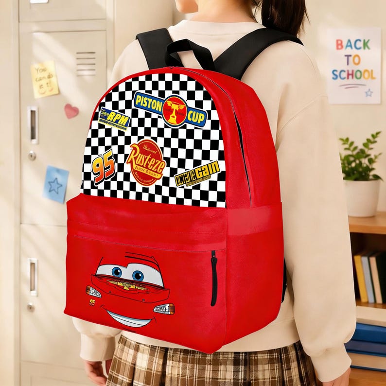 May include: Red backpack featuring Lightning McQueen from the Cars movie. The top section has a black and white chequered pattern with racing-themed patches. The backpack has a black zip and straps, ideal for carrying school supplies.