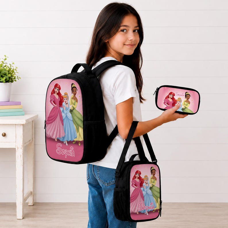 May include: A black backpack, a matching lunch bag, and a pencil case, each featuring a pink panel with illustrations of Ariel, Cinderella, and Tiana. The name Sophia is printed in white script on the pink sections of the backpack and lunch bag.