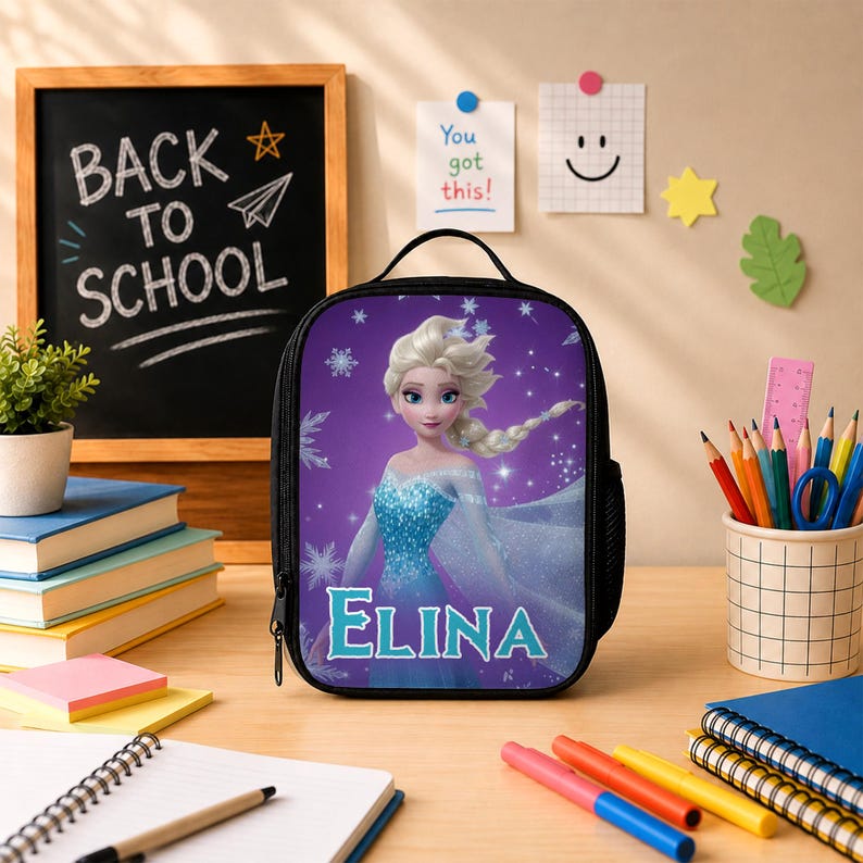 May include: A black lunch bag featuring a graphic of a blonde character in a blue dress with the name ELINA printed in blue letters. The bag has a top handle and a zipper closure, set against a purple background with white snowflake patterns.