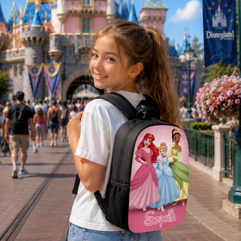May include: A black backpack featuring a graphic print of three animated princesses in pink, blue, and green dresses on a pink background. The name Sophia is printed in white script at the bottom of the graphic. The backpack has black straps.