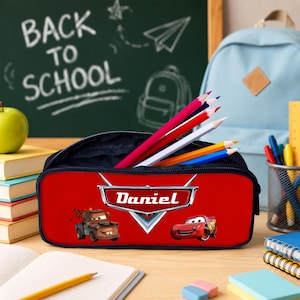 May include: A red rectangular pencil case featuring the Cars characters Mater and Lightning McQueen, with the name Daniel printed in white on a silver emblem. The case is filled with several coloured pencils and has a black zipper closure.