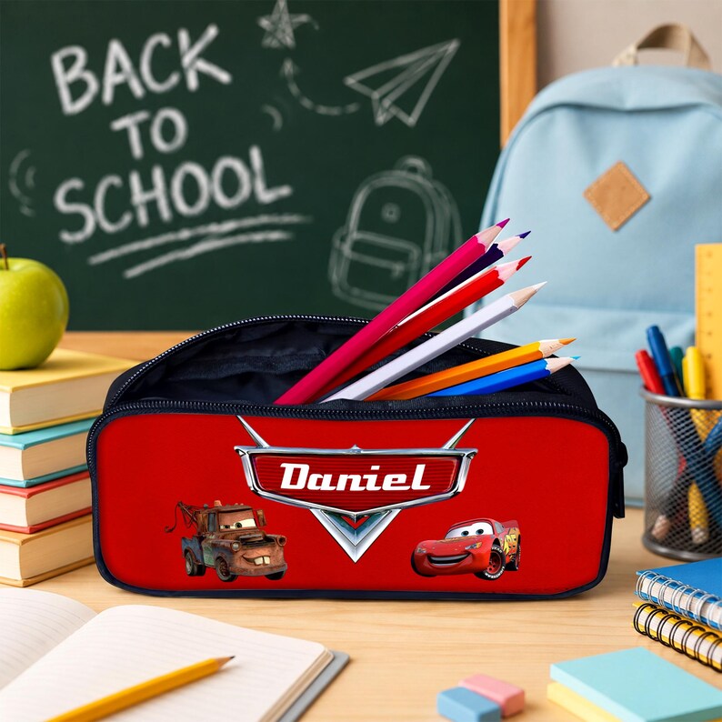 May include: A red rectangular pencil case featuring the Cars characters Mater and Lightning McQueen, with the name Daniel printed in white on a silver emblem. The case is filled with several coloured pencils and has a black zipper closure.