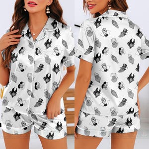 K&ouml;nnte beinhalten: Wei&szlig;es Satin-Pyjama-Set mit einem Muster aus Cartoon-B&ouml;sewicht-Gesichtern in Schwarz und Grau. Das Set beinhaltet ein Hemd mit Kn&ouml;pfen und kurzen &Auml;rmeln sowie passende Shorts. Die Kanten des Hemdes und der Shorts sind mit wei&szlig;en Paspeln versehen.