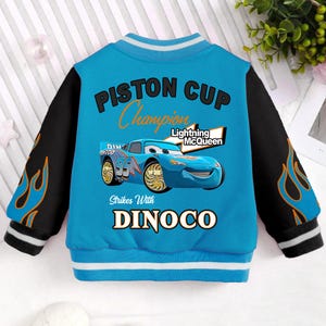 May include: Blue and black baseball jacket with a blue and orange flame design on the sleeves. The jacket has the text "Piston Cup Champion" and "Lightning McQueen" on the front. The jacket also has a picture of Lightning McQueen from the Cars movie.