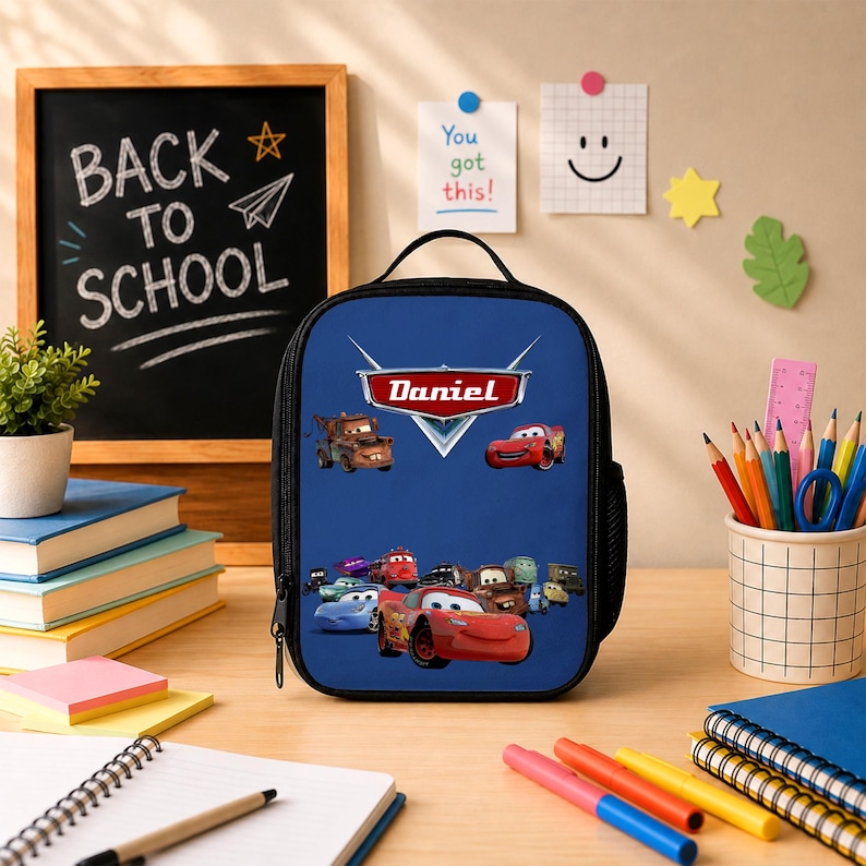 May include: A blue rectangular lunch bag featuring a graphic print of characters from the movie Cars, with the name Daniel printed in a red and white logo style. The bag has a black handle, a zipper closure, and a black mesh side pocket.