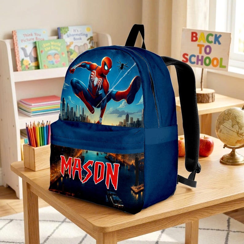 Puede incluir: Una mochila azul oscuro con un gr&aacute;fico del superh&eacute;roe Spider-Man en una pose din&aacute;mica sobre el horizonte de una ciudad. La secci&oacute;n inferior de la bolsa muestra el nombre MASON en letras rojas sobre un paisaje urbano nocturno iluminado.