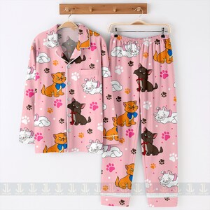 May include: Pink pajama set featuring a repeating pattern of cartoon cats, paw prints, and clouds. The long-sleeved button-up top and pants have white trim. The cats are in various colors, including orange, white, and brown, with blue and red bows.