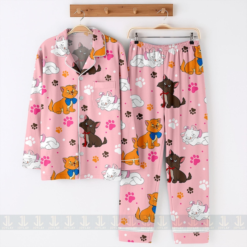 May include: Pink pajama set featuring a repeating pattern of cartoon cats, paw prints, and clouds. The long-sleeved button-up top and pants have white trim. The cats are in various colors, including orange, white, and brown, with blue and red bows.