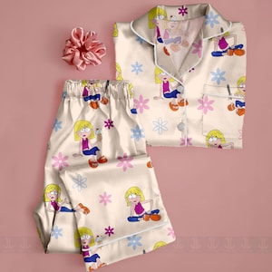 Pu&ograve; includere: Set pigiama color crema con stampa di personaggi dei cartoni animati e dettagli floreali rosa e blu. Il set include una camicia con bottoni e pantaloni abbinati. Un elastico rosa &egrave; posto vicino al pigiama.