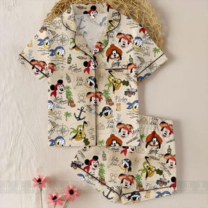 May include: A beige pajama set featuring a pirate-themed Disney character print. The set includes a short-sleeved button-up shirt and matching shorts. The print includes Mickey Mouse, Donald Duck, Goofy, and Pluto, along with anchors, treasure maps, and pirate hats.