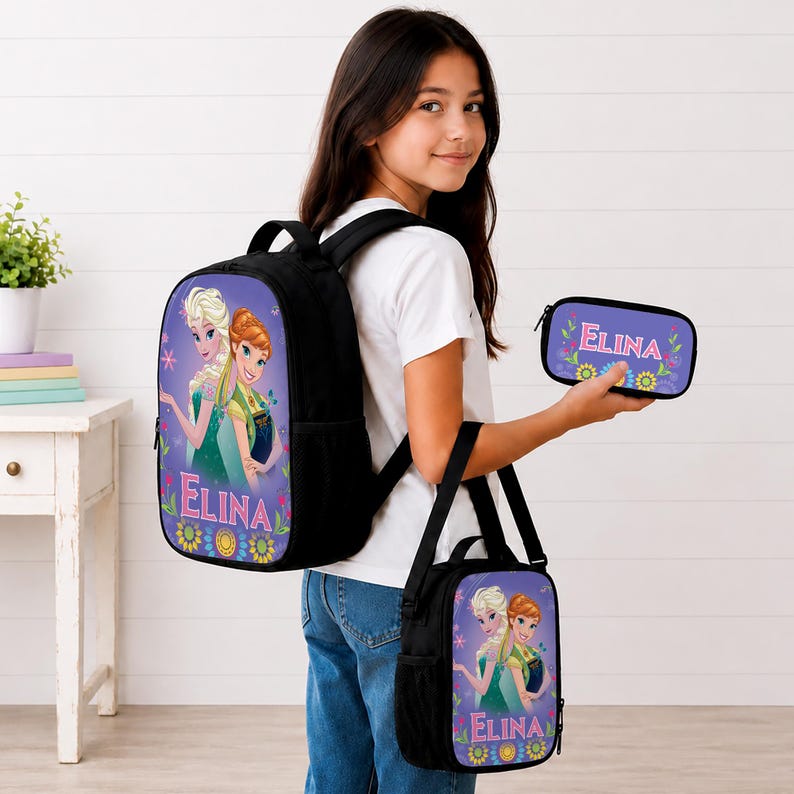 May include: A black backpack, a matching lunch bag, and a small pencil case, all featuring a purple background with a graphic of two animated characters and the name ELINA printed in pink letters above a colorful floral pattern at the bottom.