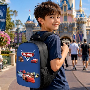 K&ouml;nnte beinhalten: Ein schwarzer Rucksack mit einer blauen Vorderseite, die einen Grafikdruck verschiedener animierter Rennwagen und den Namen Daniel in einem rot-wei&szlig;en Emblem zeigt. Die Tasche verf&uuml;gt &uuml;ber schwarze Schultergurte und seitliche Netztaschen.