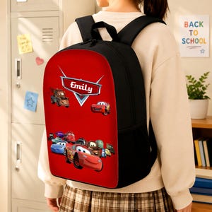 May include: A black backpack with a vibrant red front panel featuring a graphic print of various animated car characters. The design includes a central silver emblem with the name Emily printed in white text, along with multiple colourful vehicles.