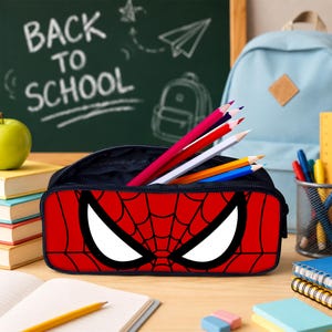 May include: A rectangular pencil case featuring a red and black spiderweb pattern with large white eye shapes on the front. The case is open, containing several colored pencils in shades of red, white, blue, and yellow, with a dark interior lining.