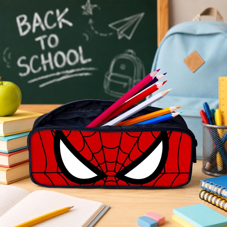 May include: A rectangular pencil case featuring a red and black spiderweb pattern with large white eye shapes on the front. The case is open, containing several colored pencils in shades of red, white, blue, and yellow, with a dark interior lining.
