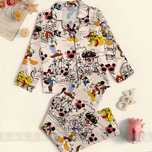 May include: A two-piece pajama set featuring a cream-colored background with a repeating pattern of cartoon pirate characters. The long-sleeved top has a button-down front and a notched collar. The matching pants have an elastic waistband.