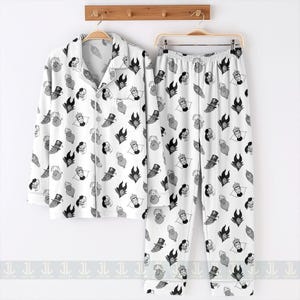 K&ouml;nnte beinhalten: Wei&szlig;es Pyjama-Set mit einem sich wiederholenden Muster aus Cartoon-B&ouml;sewicht-Gesichtern in Schwarz, Grau und Wei&szlig;. Das Set beinhaltet ein Hemd mit Kn&ouml;pfen und eine Hose, die beide an einem Holzgestell h&auml;ngen. Das Pyjama-Set ist aus weichem Material.