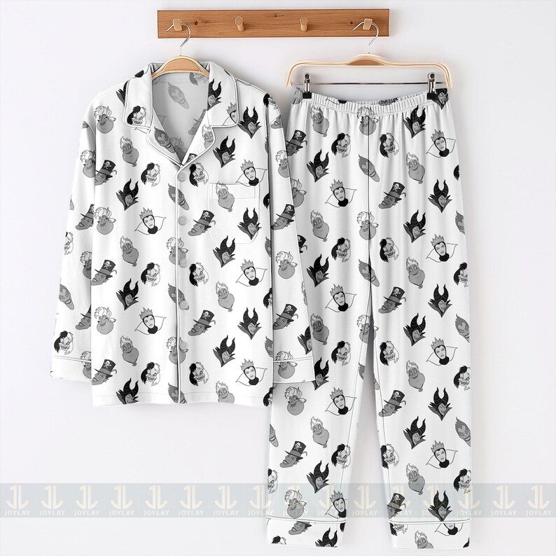 K&ouml;nnte beinhalten: Wei&szlig;es Pyjama-Set mit einem sich wiederholenden Muster aus Cartoon-B&ouml;sewicht-Gesichtern in Schwarz, Grau und Wei&szlig;. Das Set beinhaltet ein Hemd mit Kn&ouml;pfen und eine Hose, die beide an einem Holzgestell h&auml;ngen. Das Pyjama-Set ist aus weichem Material.