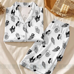 K&ouml;nnte beinhalten: Wei&szlig;es Pyjama-Set mit einem sich wiederholenden Muster von Cartoon-B&ouml;sewicht-Gesichtern in Graut&ouml;nen. Das Set beinhaltet ein Oberteil zum Kn&ouml;pfen und eine passende Hose. Der Pyjama ist aus einem weichen, bequemen Material gefertigt.