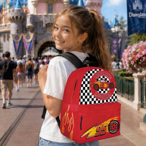 May include: Red backpack featuring Lightning McQueen from the Cars movie. The backpack has a checkered pattern, flames, and the number 95. The design includes the text "Lightning McQueen" and a tire graphic. The backpack is worn by a person.