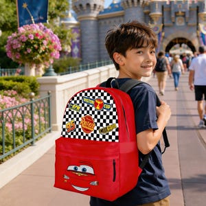 May include: A red backpack with a Lightning McQueen design from the Cars film. The top flap features racing patches and a chequered flag pattern. The backpack is worn by a person. The bag has a large front pocket.
