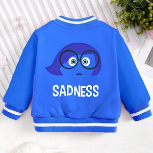 May include: Blue and white baseball style jacket with a cartoon character of a blue female character with glasses and the word "SADNESS" printed on the back.