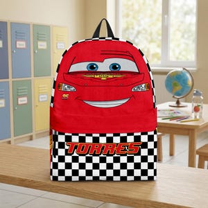 Peut inclure: Sac &agrave; dos rouge avec un visage de voiture de dessin anim&eacute;, des yeux bleus et une bouche souriante. La partie inf&eacute;rieure pr&eacute;sente un motif &agrave; carreaux noir et blanc avec le nom "TORRES" en rouge. Il a une poign&eacute;e noire.