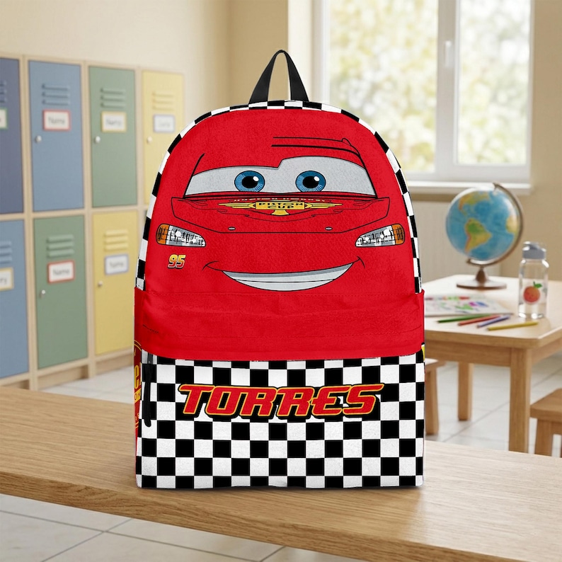 Peut inclure: Sac &agrave; dos rouge avec un visage de voiture de dessin anim&eacute;, des yeux bleus et une bouche souriante. La partie inf&eacute;rieure pr&eacute;sente un motif &agrave; carreaux noir et blanc avec le nom "TORRES" en rouge. Il a une poign&eacute;e noire.