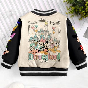 May include: Cream-colored bomber jacket with black sleeves and trim. The back features a Disneyland graphic with Mickey and Minnie Mouse, Donald Duck, and other characters. The text "Disneyland - Est. 1955" is also visible.