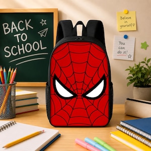 May include: A black backpack featuring a prominent red front panel with a black spiderweb pattern and large white eyes. The design mimics a superhero mask, with black shoulder straps and side mesh pockets, positioned on a wooden desk surface.