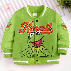 May include: A green bomber jacket featuring a large graphic of Kermit the Frog with the word "Kermit" in orange script. The jacket has white and black accents, including stripes on the collar, cuffs, and hem. Two small Kermit graphics are on the sleeves.