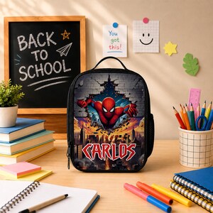 May include: A black lunch bag featuring a graphic of a red and blue superhero bursting through a brick wall above a city skyline. The name CARLOS is printed in bold red letters at the bottom. The bag has a top handle and a side mesh pocket.