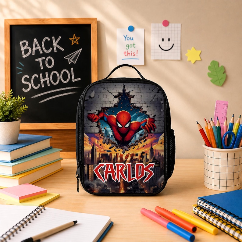 May include: A black lunch bag featuring a graphic of a red and blue superhero bursting through a brick wall above a city skyline. The name CARLOS is printed in bold red letters at the bottom. The bag has a top handle and a side mesh pocket.