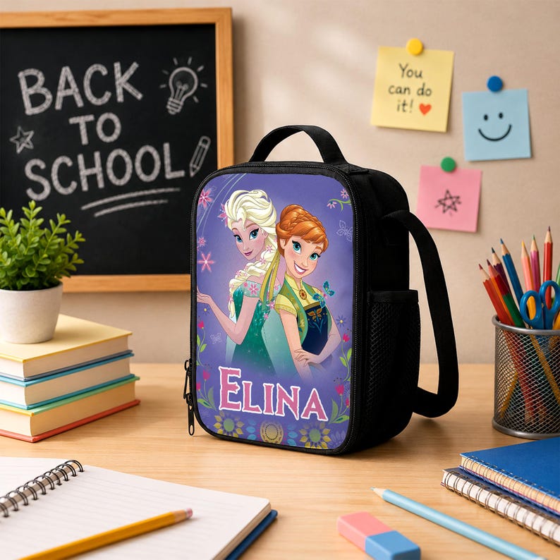May include: A black rectangular lunch bag featuring a graphic print of two animated characters with blonde and auburn hair, surrounded by floral accents and the name ELINA in large white letters. The bag has a top handle and a side mesh pocket.