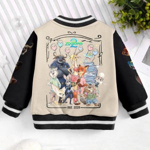 May include: A beige and black varsity jacket featuring characters from the animated film "Zootopia 2". The jacket has black sleeves and a beige body with a graphic of the film's characters. The jacket has a black and white striped collar and cuffs. The text "Zootopia 2" is visible.