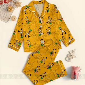 May include: Mustard yellow pajama set featuring an all-over print of the Disney character Pluto. The long-sleeved top and pants are trimmed with white piping. The top has a button-down front and a notched collar. The pajamas are made of a silky material.
