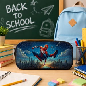 Puede incluir: Un estuche rectangular con un estampado gr&aacute;fico de Spider-Man en una pose din&aacute;mica sobre el horizonte de una ciudad. El estuche tiene una cremallera negra y un marco oscuro, mostrando una ilustraci&oacute;n detallada del personaje bajo un cielo azul.