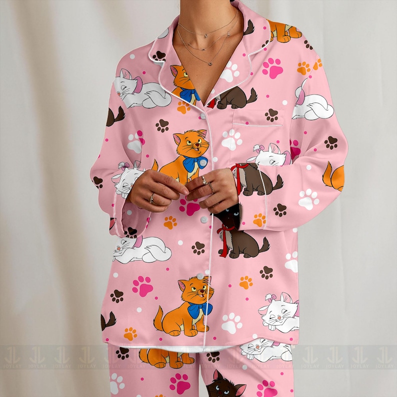May include: Pink pajama set featuring a repeating pattern of cartoon kittens, paw prints, and clouds. The long-sleeved top has white trim and a button-down front. The pajama set is made of a soft, comfortable material.
